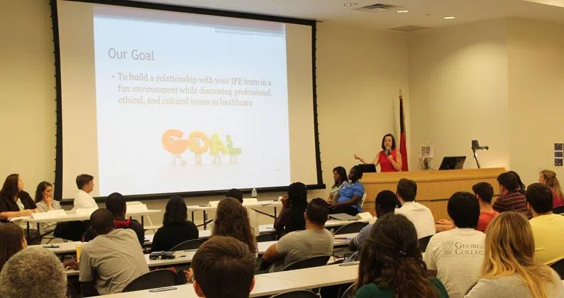 Instructor stands at the front of a room with a microphone talking about the slide shown on the presentation screen. Students sit at tables in front of her. 