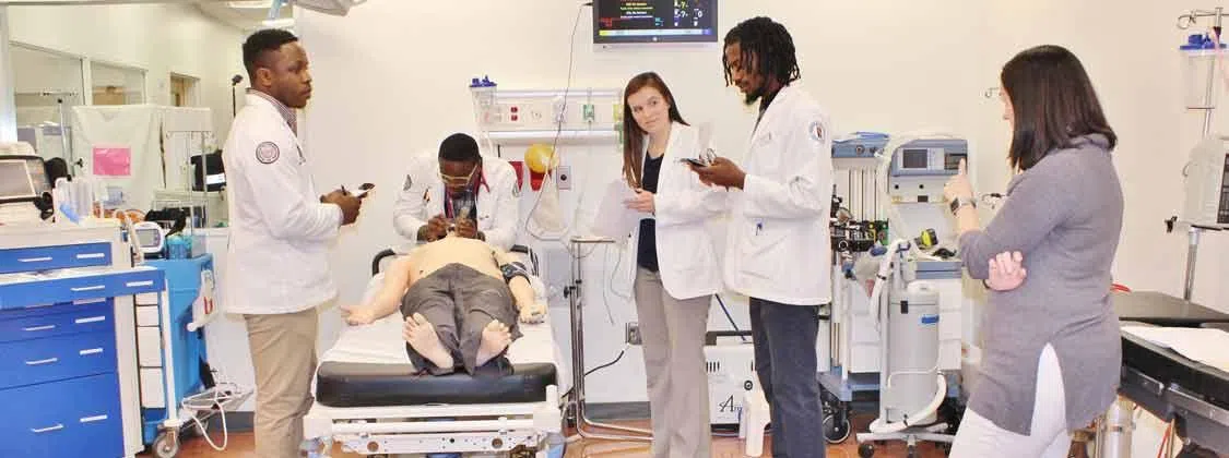 Four students in medical coats examine a manikin patient while an instructor stands by. 