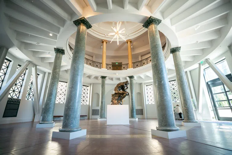 Large bronze statue of "The Thinker" sitting in the middle of a round museum rotunda with marble columns