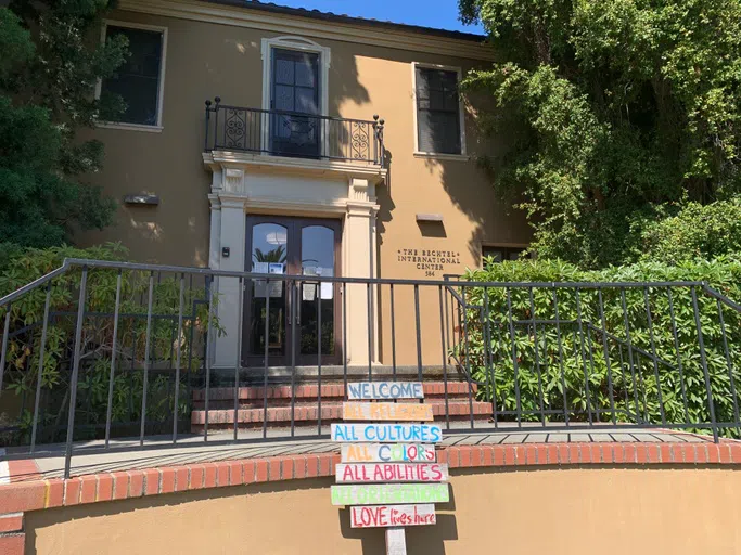 Facade of two-story building with balcony, stairwell, an entrance surrounded by trees, and a hand-painted sign indicating welcome to those of all religions, cultures, colors, and abilities