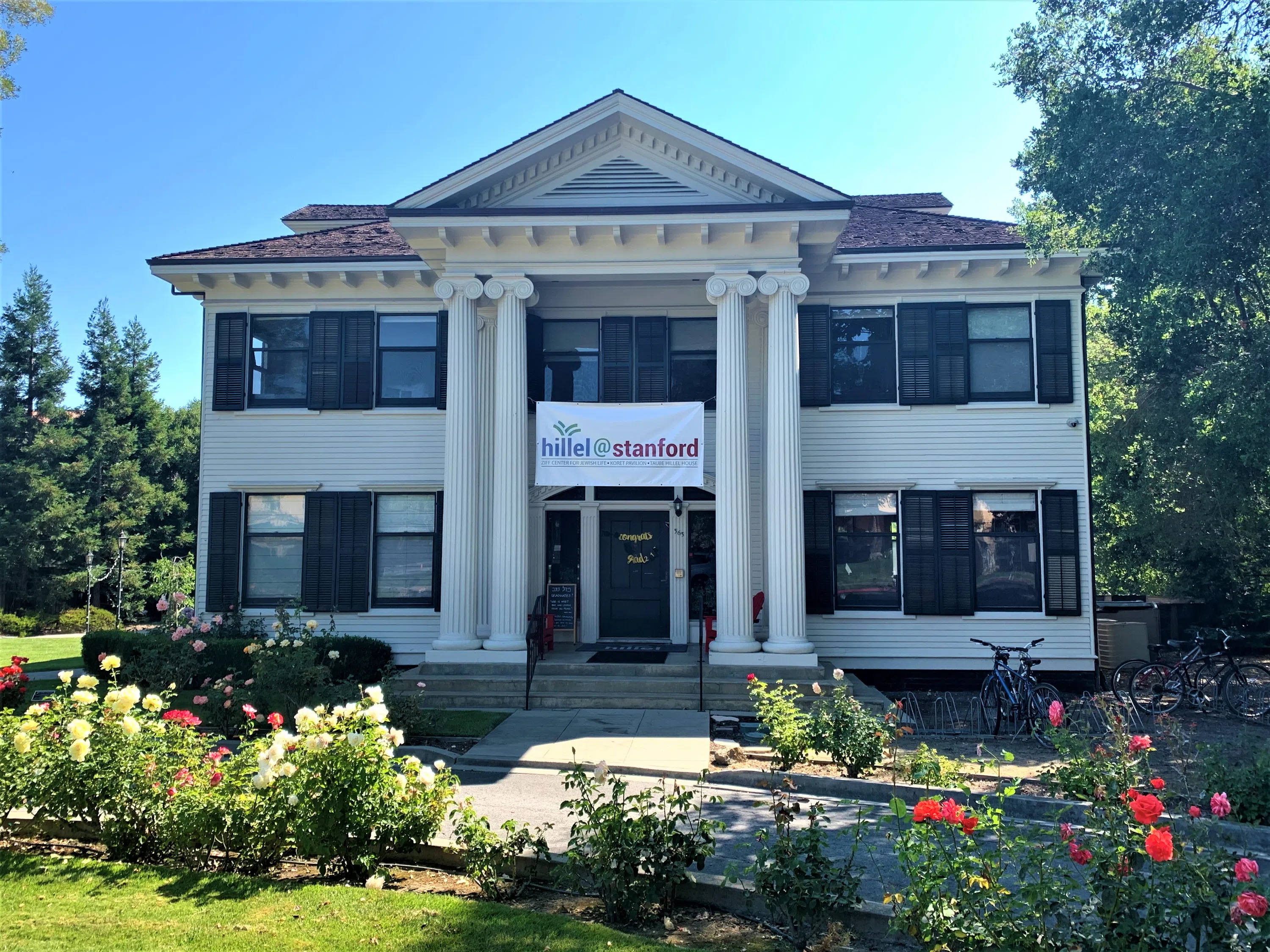 Two-story house with white columns, a "Hillel at Stanford" banner, and floral landscaping.