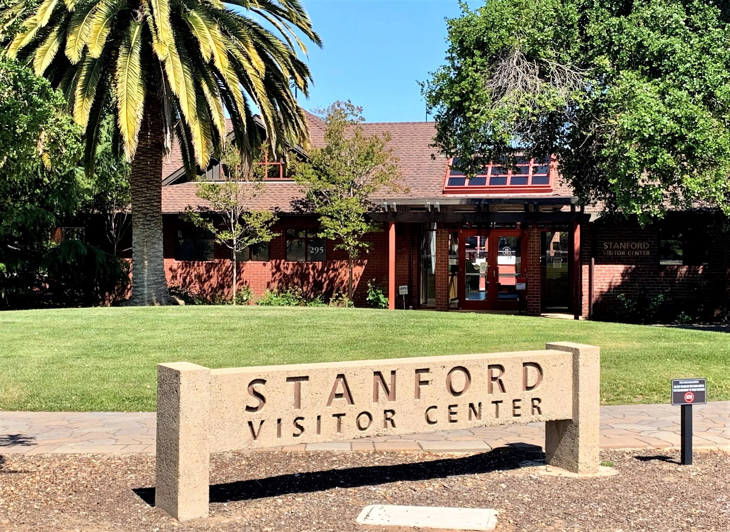 Stone sign reading "Stanford Visitor Center" in front of a single-storyBrick building with a palm tree and grassy hill in between.