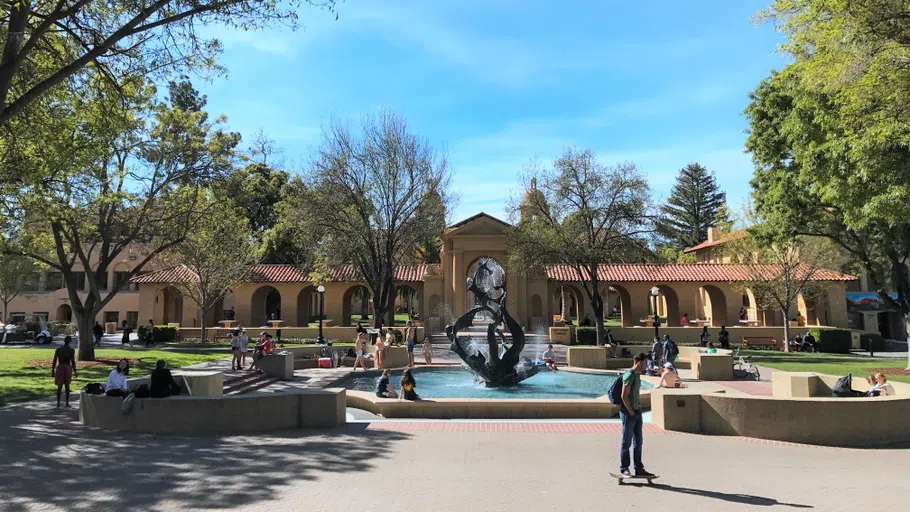 Students and bicyclists around a splashing fountain containing a large claw-like sculpture, situated in a plaza in front of mission-style building.