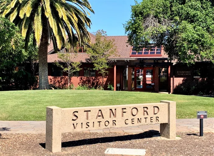 Stone sign reading "Stanford Visitor Center" in front of a single-storyBrick building with a palm tree and grassy hill in between.