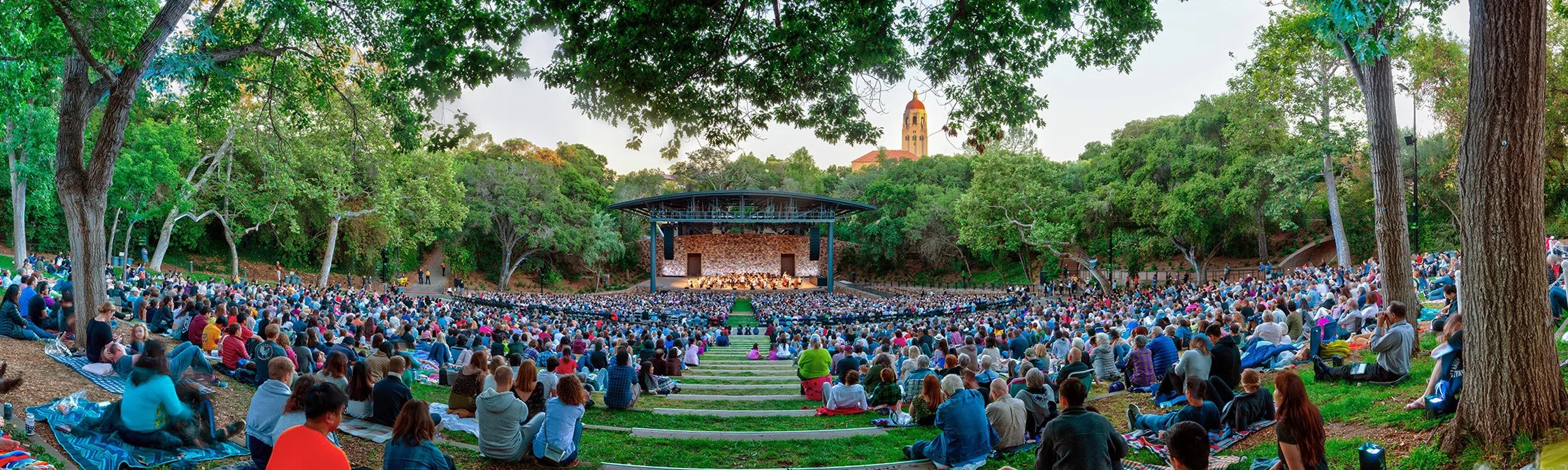 Hundreds of people seated on terraced lawns leading down to a modern covered stage in a tree-lined amphitheater