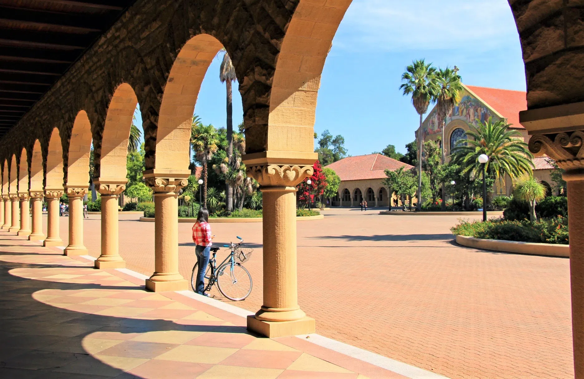 A student stands with bicycle between two of the many sandstone arches that surround a mission-style, paved courtard containing multiple "oases" of trees.