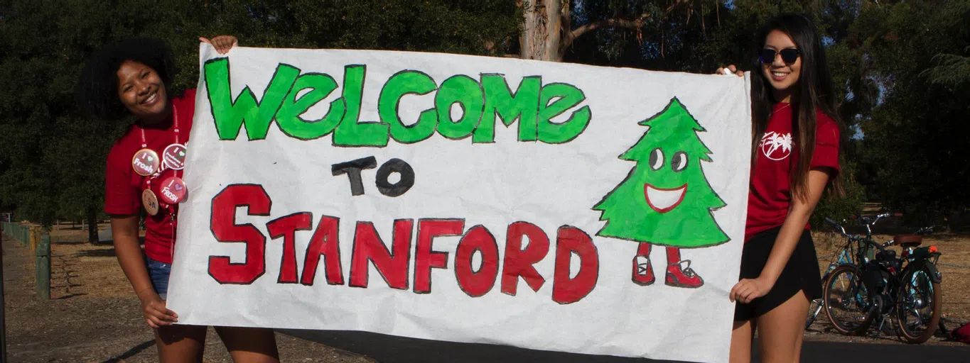 Two students hold up a painted sign between them. The sign reads, "Welcome to Stanford" in red and green block letters, and has a painting of a Stanford tree