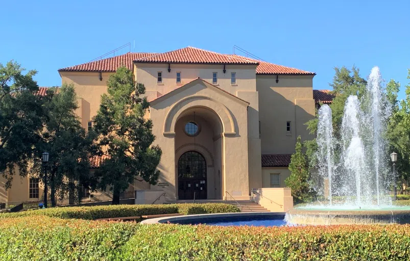 Exterior of large sandstone auditorium with tall archway at facade and fountain shooting up water in front.