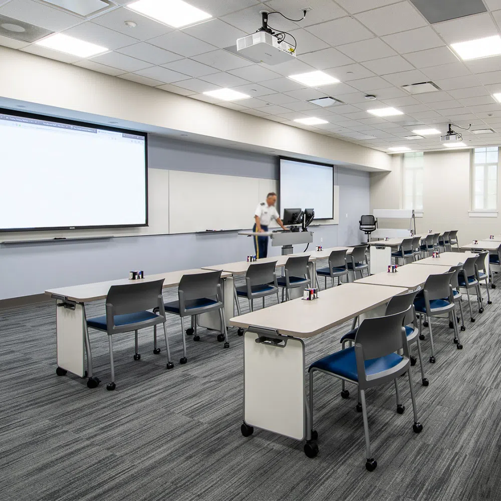 Rows of tables with chairs in Bond Hall classroom. Person stands in the front of the space behind a podium and in front of two projection screens. 