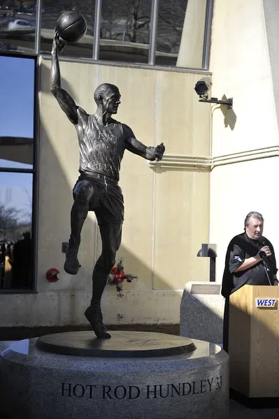 Image shows Men's Basketball Head Coach, Bob Huggins, speaking in front of "Hot Rod" Hundley Statue by entrance to the WVU Coliseum.