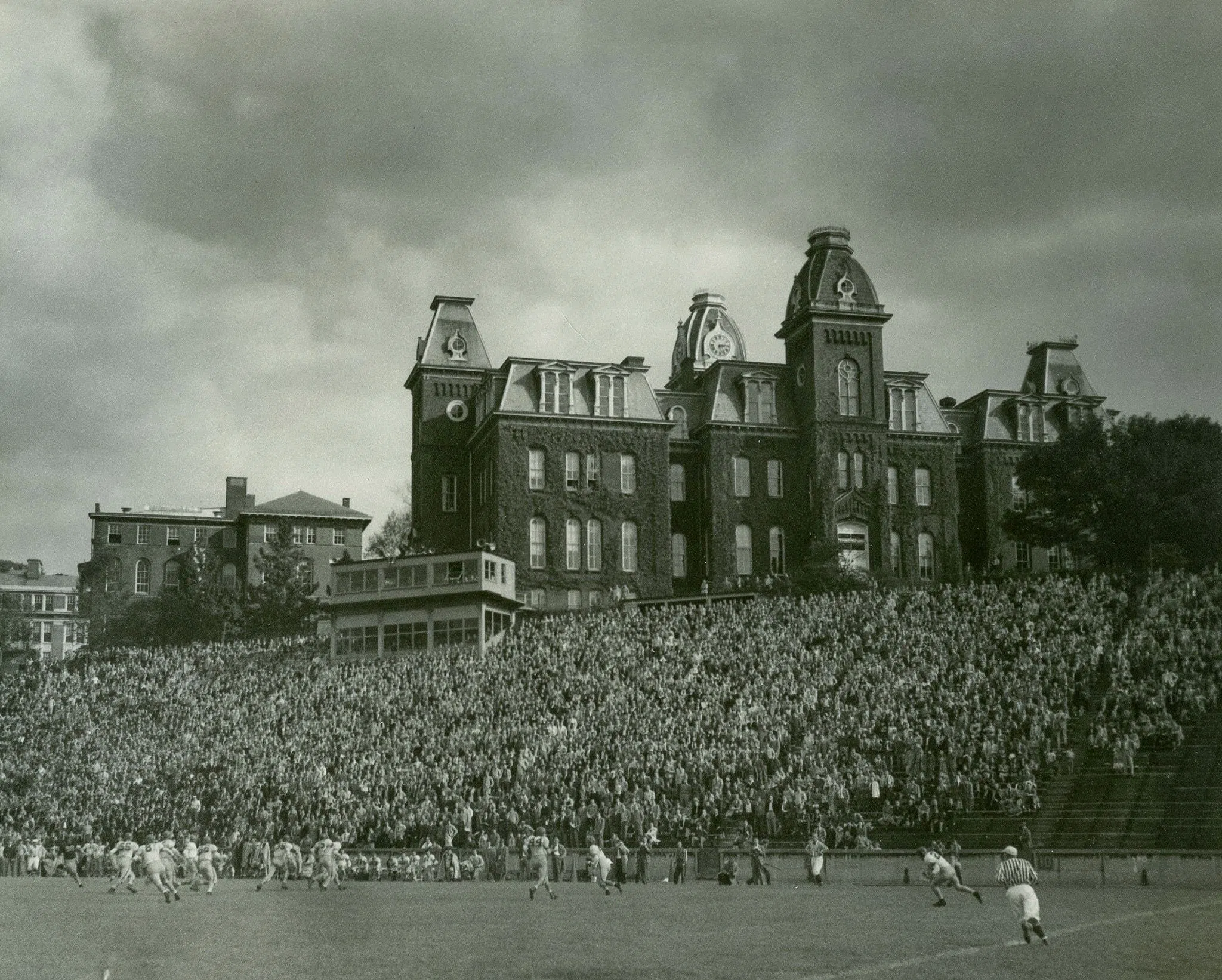 Black and white image captures football game at Old Mountaineer Field with Woodburn Hall, covered in ivy, at the top of the stadium.