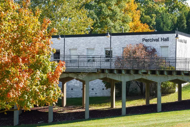 Image shows the exterior of Percival Hall, a gray brick building with an elevated pedestrian bridge in front.. 