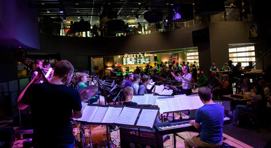 The One O'Clock Band, a jazz band at UNT, plays in a large space in the University Union building called the Syndicate. People sit around the stage, listening to the performance.