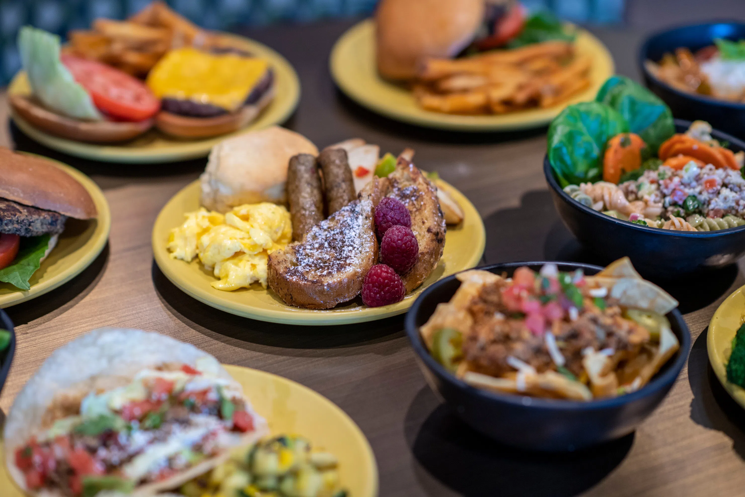 Close up of a breakfast plate holding french toast, eggs, sausage and a biscuit. Other plates of food surround the breakfast plate and are blurred in the image. 