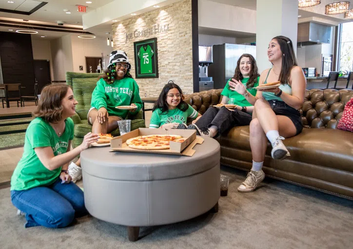 5 students sit on couches and on the floor of a common area. All of the students are chatting and smiling. A pizza sits in the middle of them on a circular ottoman