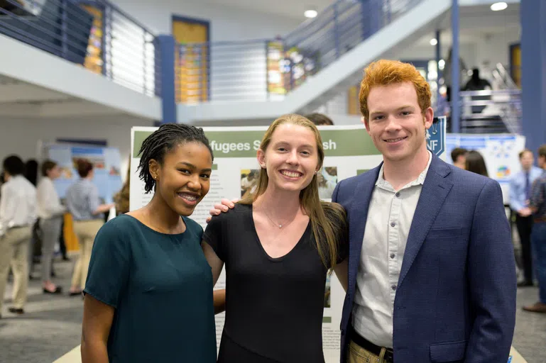 Three students stand in front of a research poster, smiling for the camera