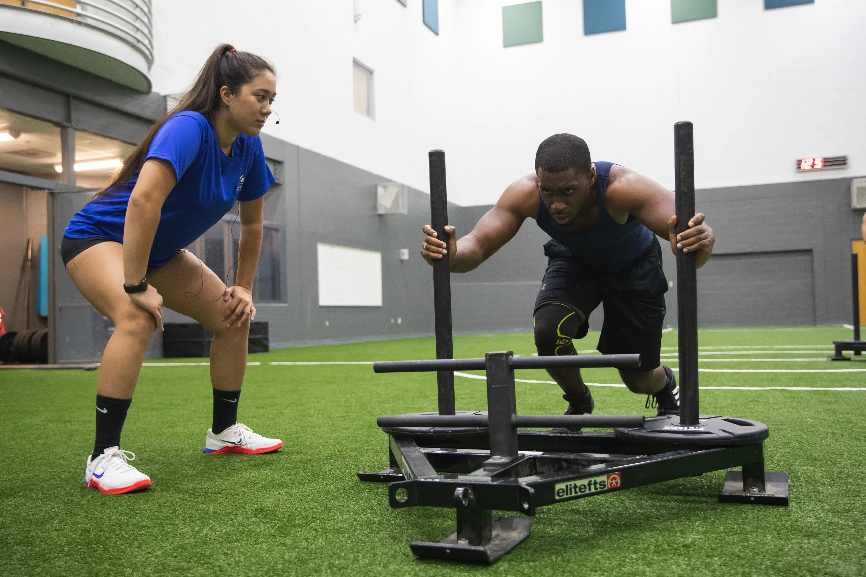 A female student coaches a male student as he trains on a piece of sled equipment in the HLS building.