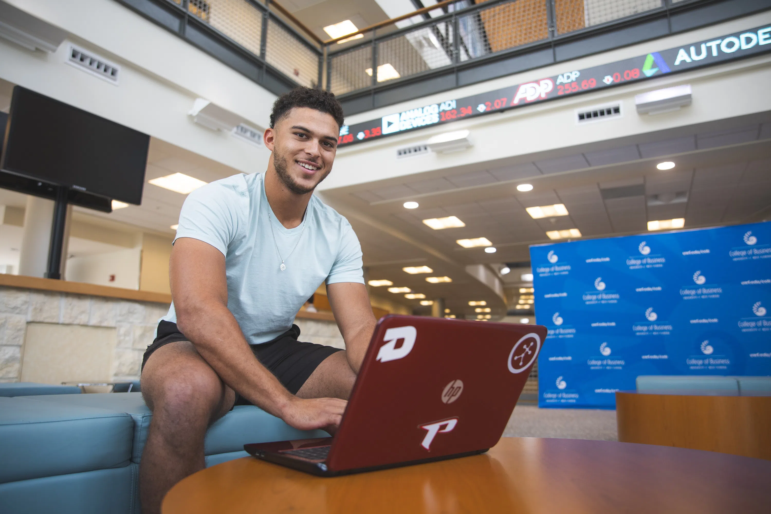 Student on laptop in the College of Business Atrium with the New York Stock Exchange Ticker in the background. 