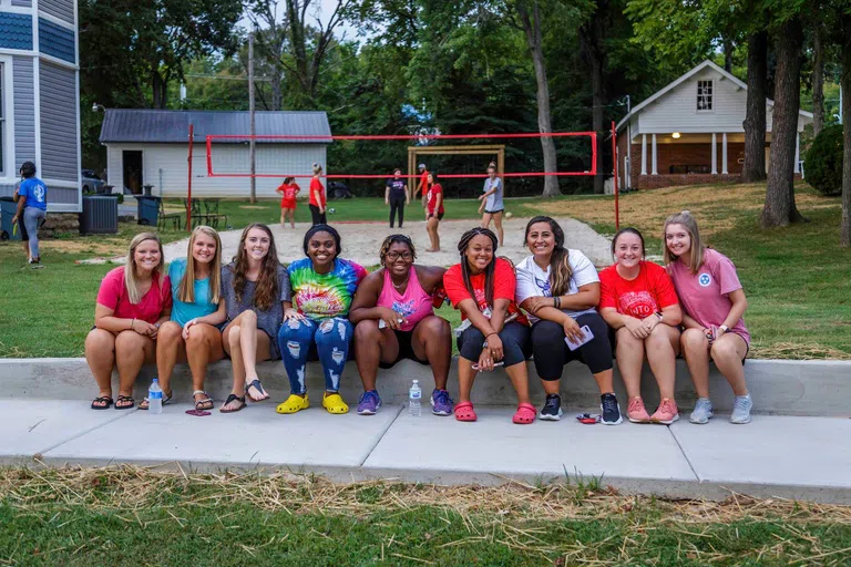 Students sitting outside of the Student Union in front of a volleyball game.