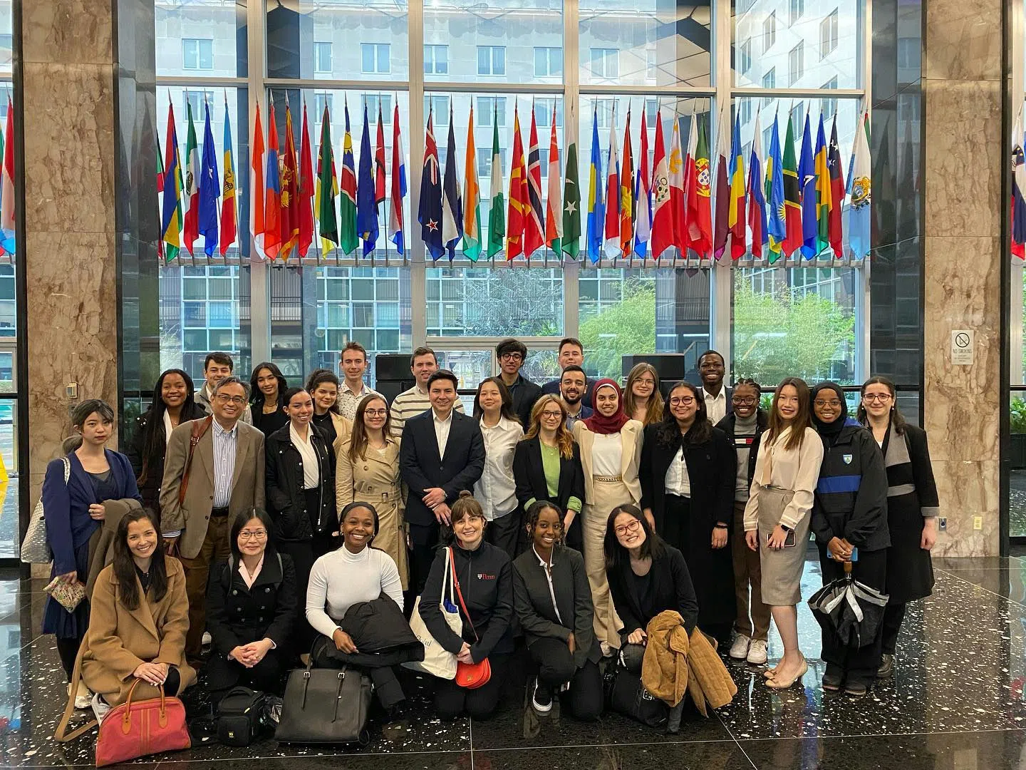 Over 30 formally dressed Penn representatives, some standing and others sitting, pose for a group photo in a dignified lobby beneath various country flags.