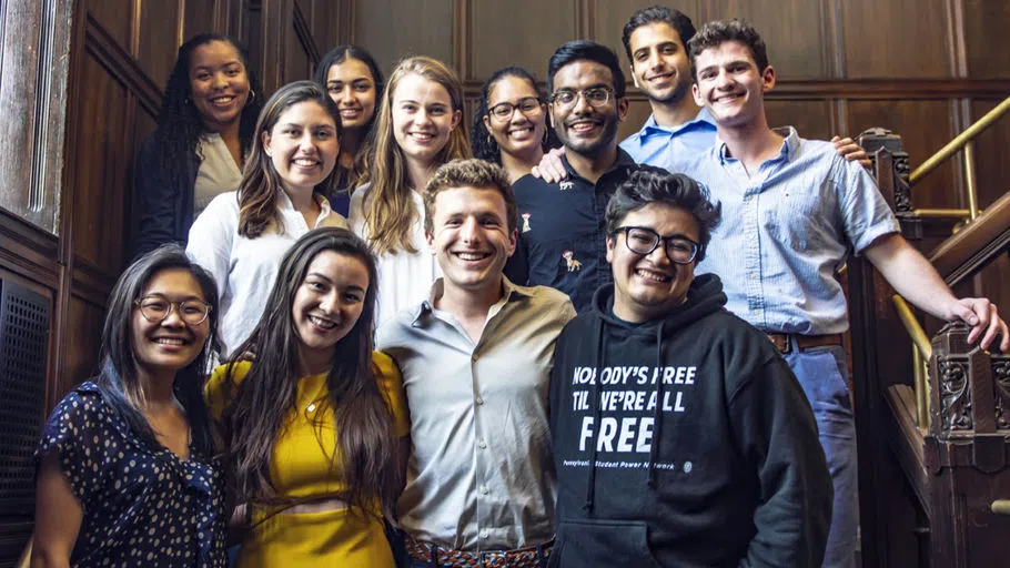 Twelve visually diverse Civic Scholars stand together, hands on each other's shoulders, smiling. One student wears a sweatshirt with the message: "Nobody's Free Until We Are All Free."