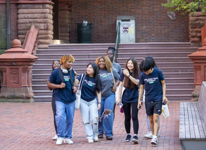 A casual group of students strolling by Fisher Fine Arts Library's red sandstone steps, some with tote bags, water bottles, and one wearing headphones.