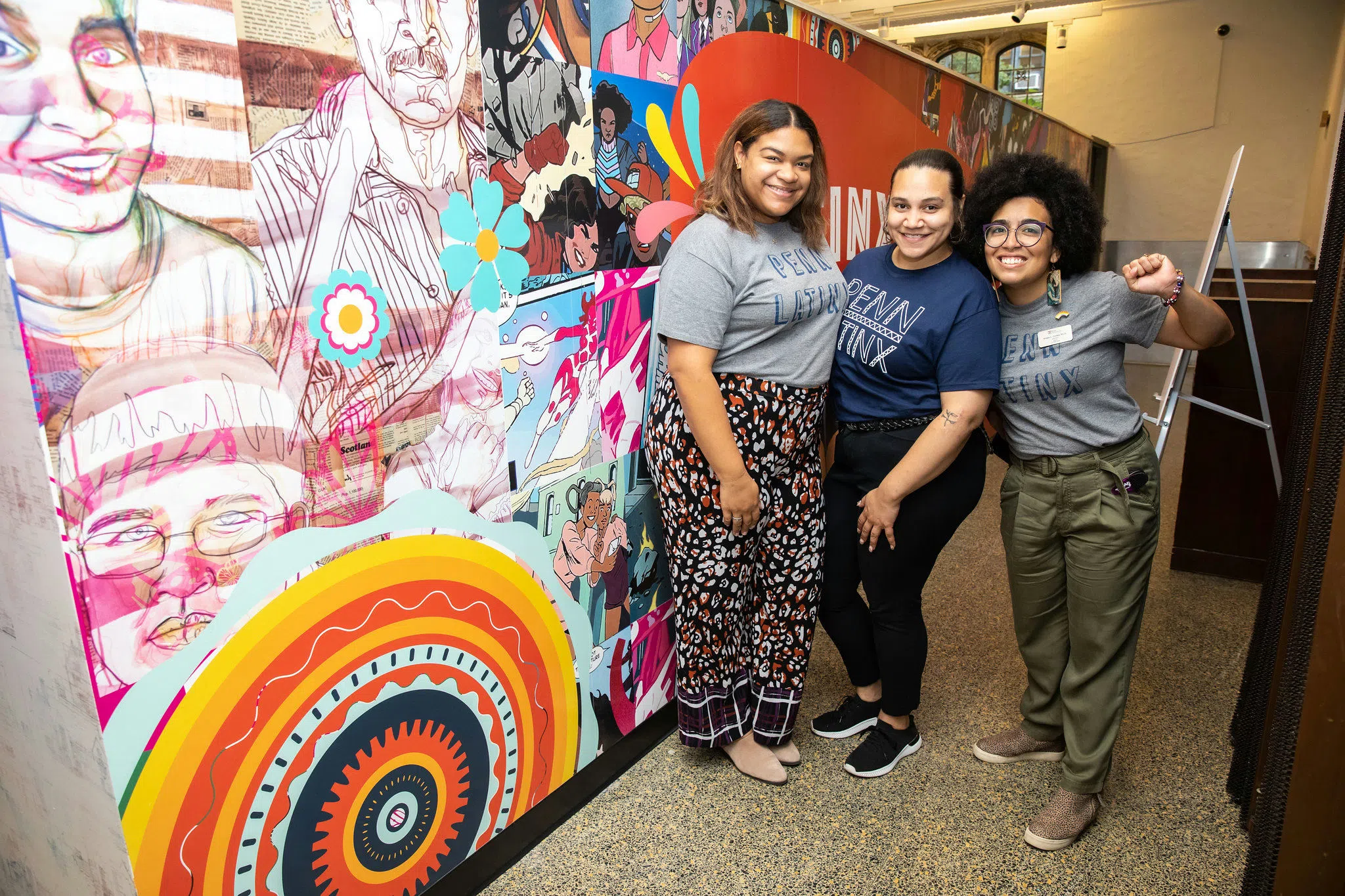 Three women are standing together with grey "Penn Latinx" shirts in front of a mural in the ARCH building. One is holding their fist up as a gesture of solidarity and pride. 