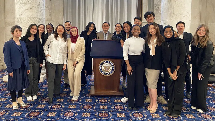 Twenty students and staff pose on the floor of the United States Senate, with the official seal prominently displayed on a podium.