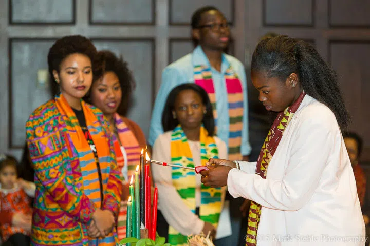 A student lights one of seven green, black, and red candles. Four additional students are in the background and they are all wearing kente scarves of various colors.