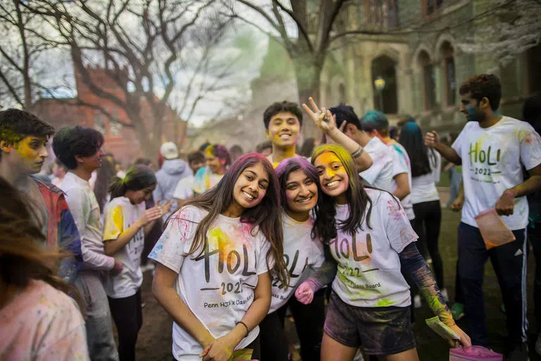 Four students, three with long hair smiling and one making a peace sign, wearing "Holi 2022" white shirts, covered in colorful chalk powder.