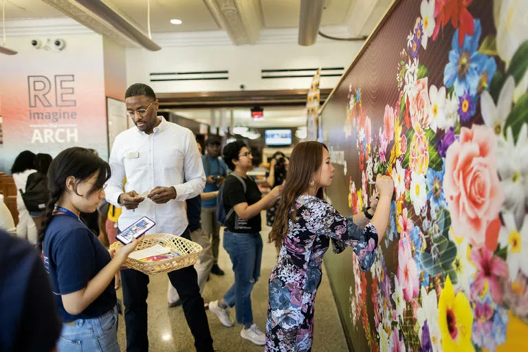 Staff member adding writing to a mural depicting different varieties of flowers. In the background, there is text on a wall that reads "Re-Imagine ARCH". 