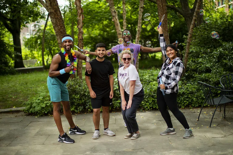 A staff member in a flower necklace, green shorts, and headband poses joyfully with students. One student blows bubbles, capturing their happiness.