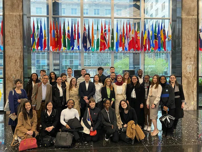 Over 30 formally dressed Penn representatives, some standing and others sitting, pose for a group photo in a dignified lobby beneath various country flags.