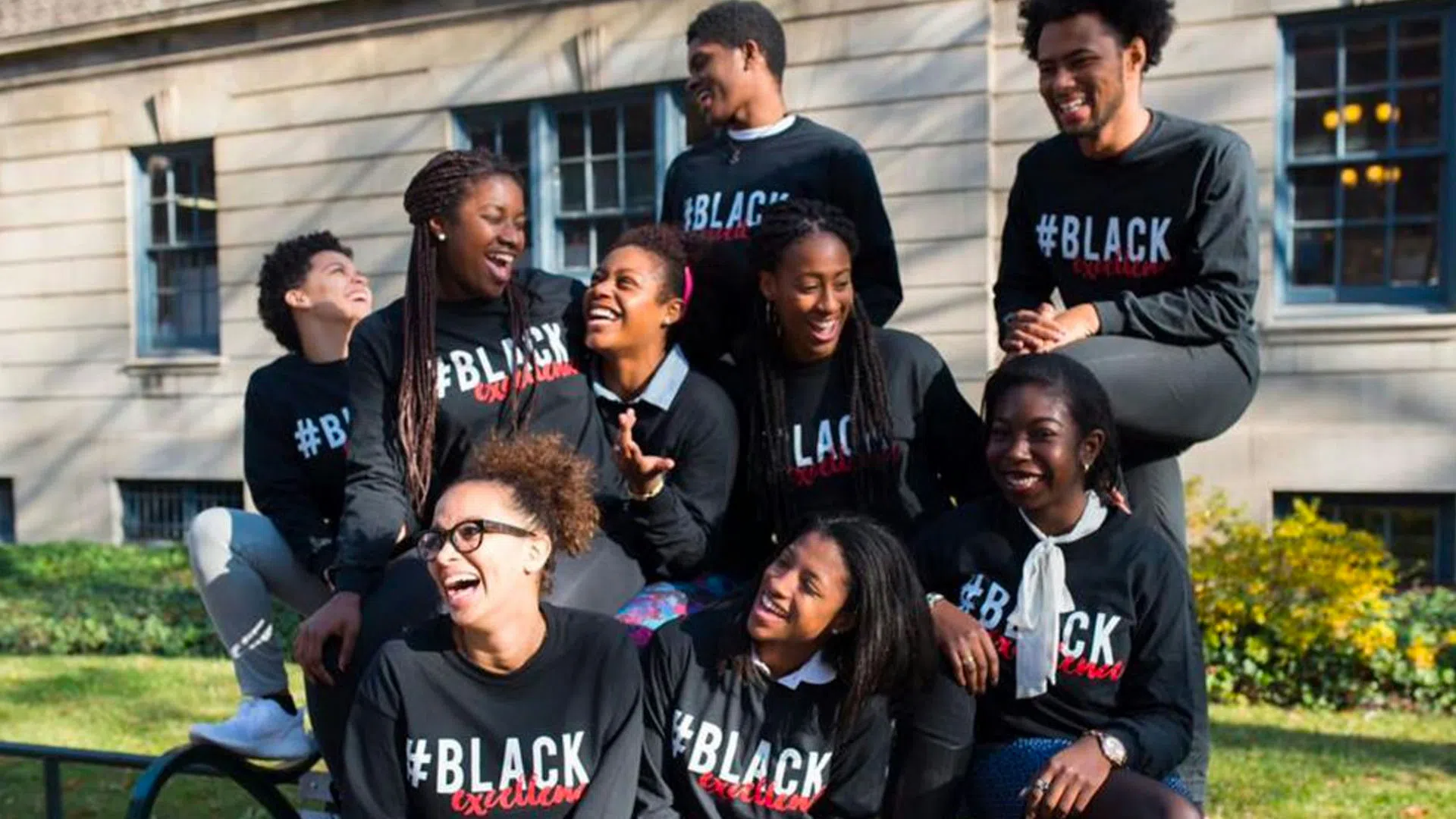 Nine students are outside together laughing. They all are wearing the same black sweatshirt with red and white text. The text reads "# Black Excellence".