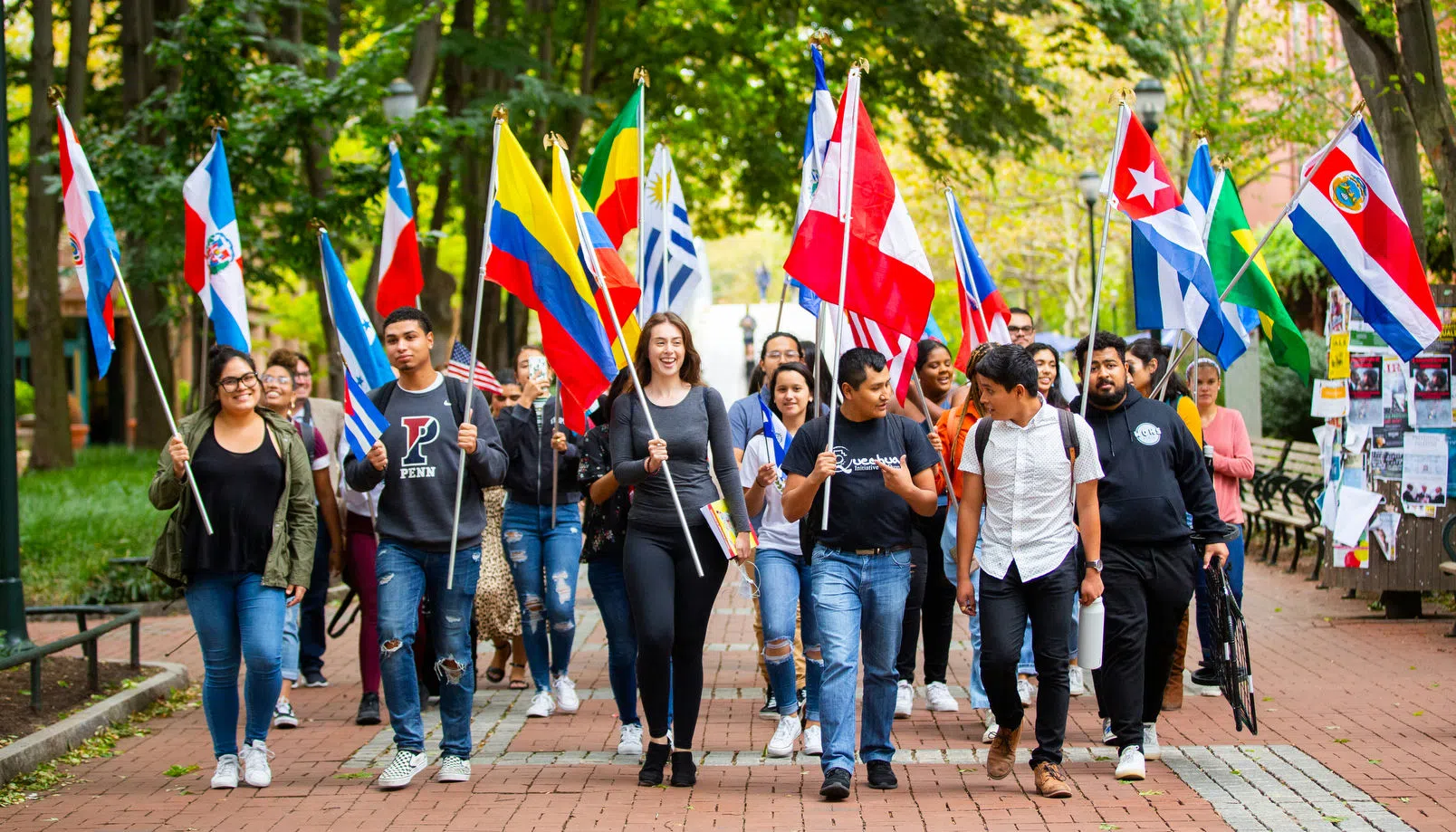 Group of 20 Penn students and community members walking down Locust Walk holding flags representing various Latin American countries.