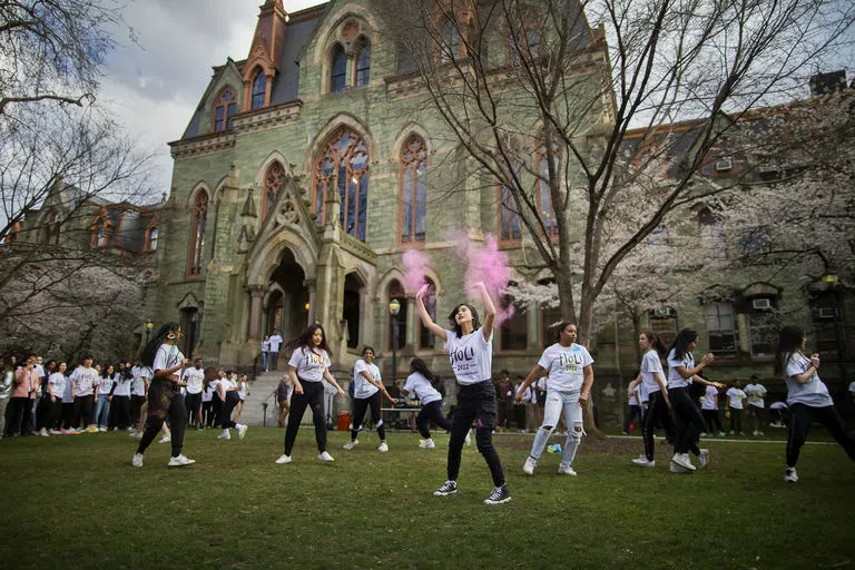 A crowd of excited students before College Hall, one in the center with raised hands, pink powder swirling around them.