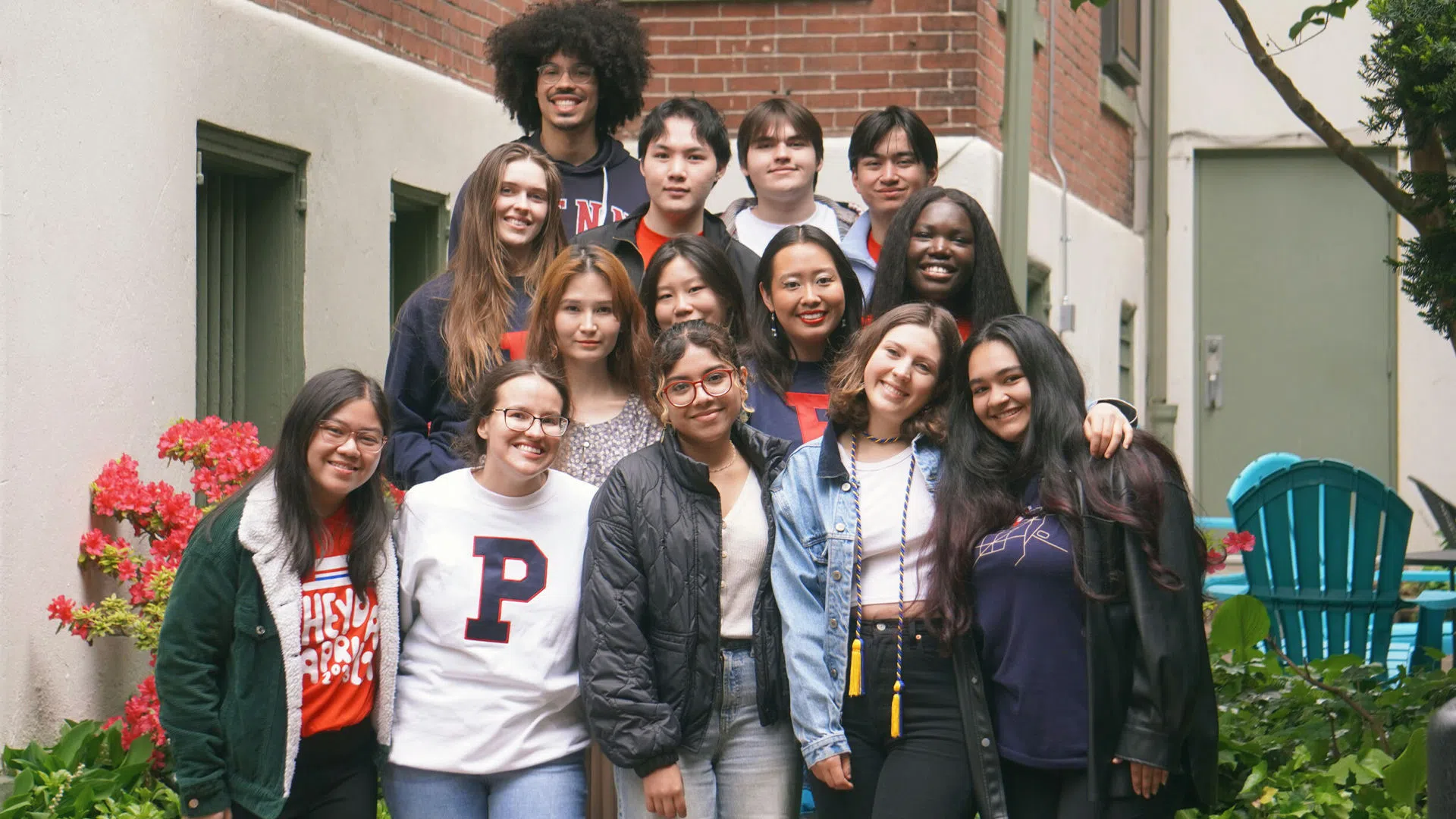 Fourteen visually diverse students gather beside a beige house with brick and green details, many sporting red and blue Penn shirts and sweaters.