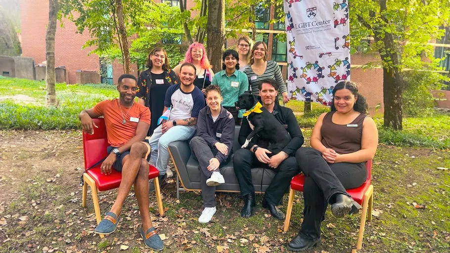 Center staff and student assistants pose for a photo, some seated, some standing. A staff member holds a dog, and they all wear pronoun nametags. One staff's shirt reads "You Belong."