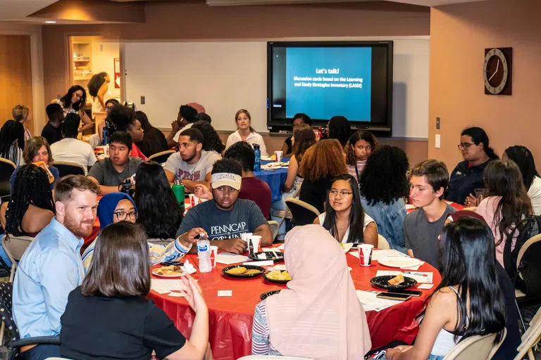 Dozens of students gather around round tables, conversing and eating at a workshop, with a TV in the background reading "Let's Talk."