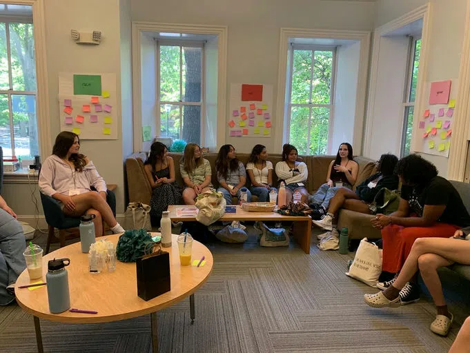 Ten students sit on couches, fully engaged in a workshop, with posters in the background that display "age," "race," and "gender" along with responses written on sticky notes.