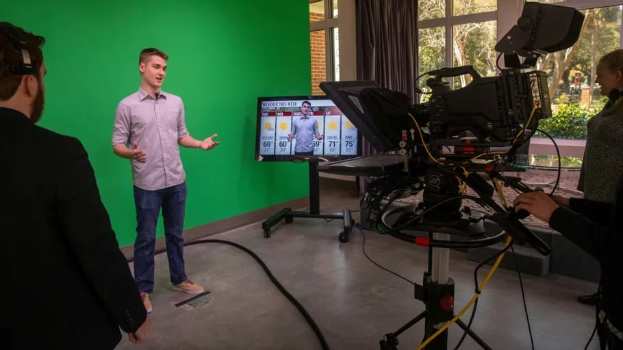 A student producer and camera operator help another student give a weather report in front of a green screen in the Kennedy Greenhouse Studio.