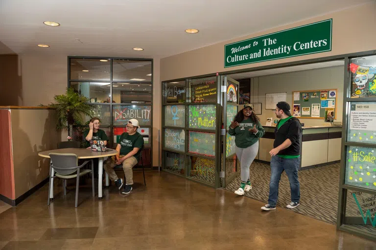 The entrance to the Culture and Identity Centers in Millett Hall. Two students walk out of the centers talking and two students sit at a table outside the centers talking.