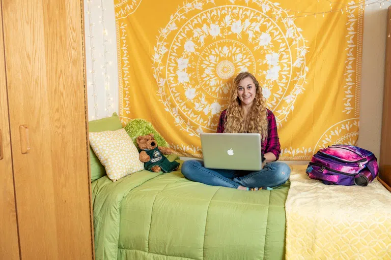 A student sits on dorm bed while working on laptop.