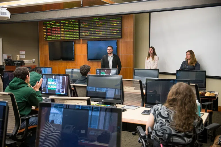 Students sit at the desk in the Soin Trading Lab and listen to a presentation. 