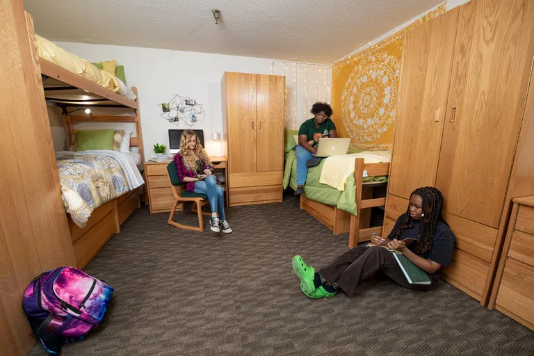 Three students are sitting in studying in their triple dorm room. Featured is one bunked bed and a single bed, wardrobes, dresser, and desk. 