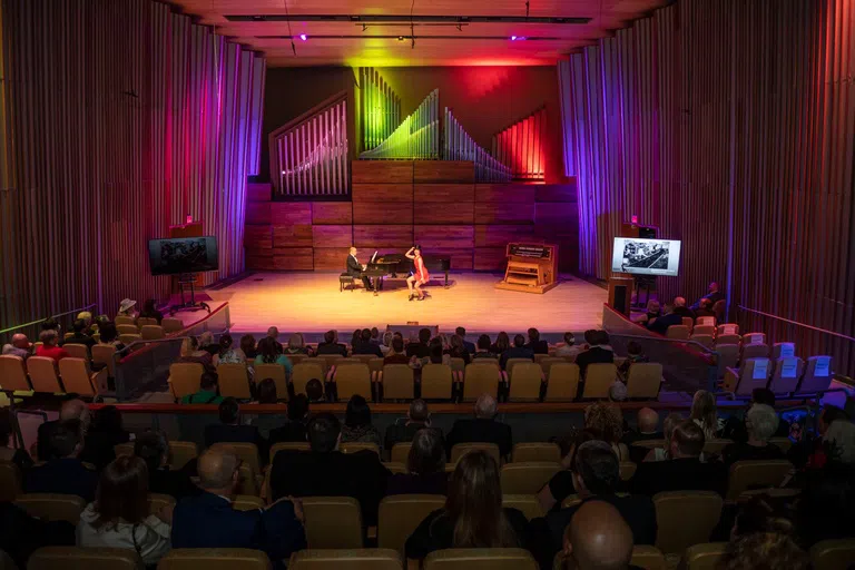 A person plays the piano while another person dances and sings on stage in the Benjamin & Marian Schuster Hall. An audience of people sit in the hall watching the performance.