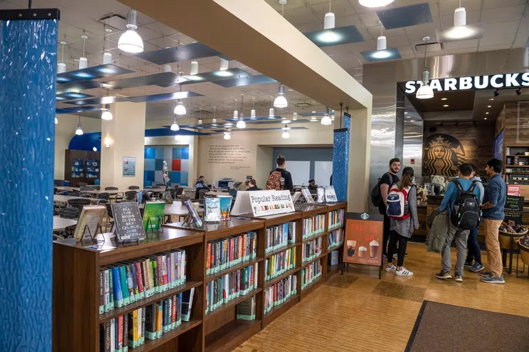 Students stand in line at the Starbucks in the library. Various tables for seating and a bookshelf with popular books are also in the frame. 