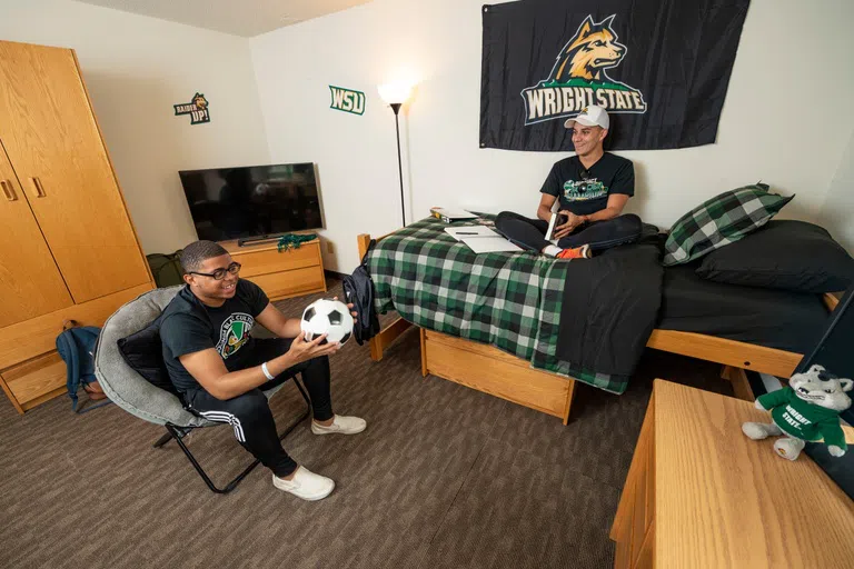 Two students sit in a dorm room, one student sits on the bed with a book and notebook out, the other student sits in a chair holding a soccer ball. In the photo, there is featured a bed, dresser, wardrobe, tv stand, desk, and wright State decorations.