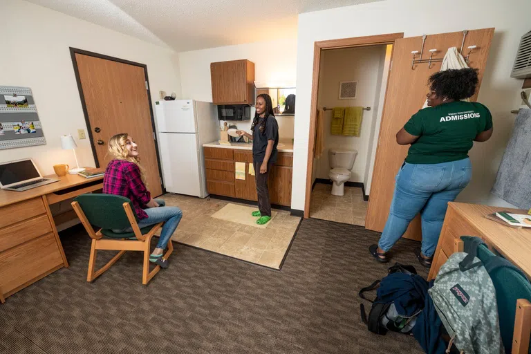 Three students are chatting in their triple dorm room. Featured in the photo is a private bathroom, small kitchen with a regular sized fridge, and some of the dorm furniture.