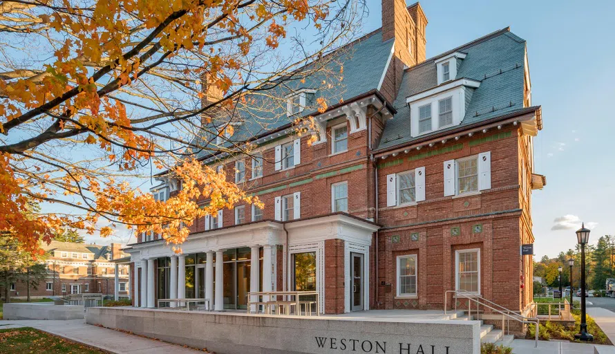 An image of Weston Hall, a red brick building with windows and a slanted roof. Yellow leaves from a tree dip into the photo from the left side, and the bottom shows the slab by the walkway that reads "WESTON HALL"